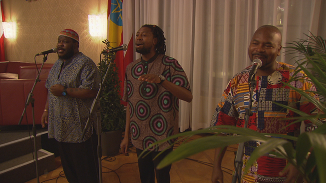 3 Männer in T-Shirts aus traditionellen Stoffen singen gefühlvoll an drei Mikrofonen 