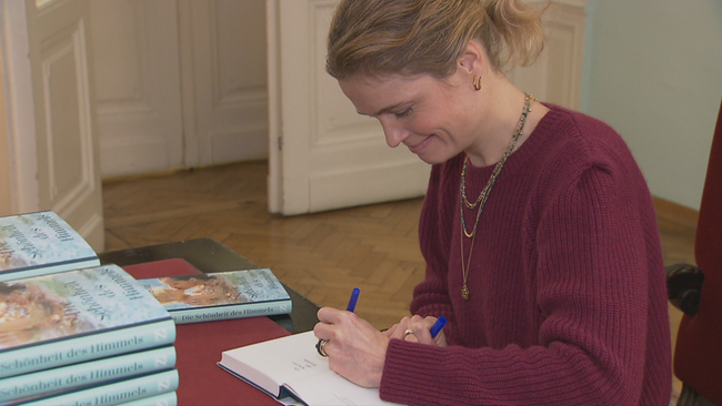 Sarah Biasin in weinrotem Pullover mit zartem Goldschmuck, lächelnd, signiert Bücher in blauem Band