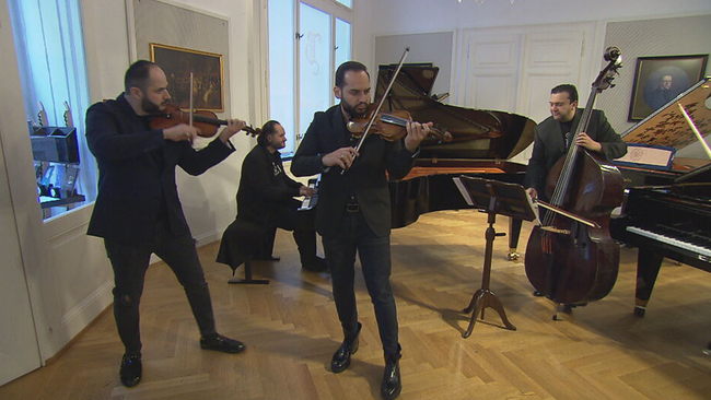 4 Männer in schwarzen Anzügen, hinten einer am Flügel und einer am Kontrabass, vorne zwei an den Geigen