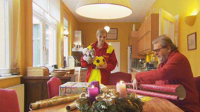 Steinhauer beim Geschenke einpacken in einer gelb und orange gestrichenen Wohnküche. Die Kerzen am Adventskrank brennen, Mitarbeiterin vom Samariterbund bringt Geschenke