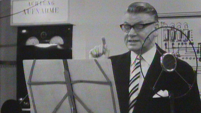 Schwarz/weiß Aufnahme Conrads während einer Radioaufnahme moderierend mit erhobenem Zeigefinger