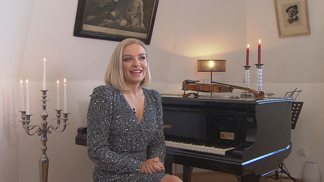 Lidia Baich in glitzerndem Kleid am Klavierstuhl. Im Hintergrund klassische Kunst, ihre Geige am Klavier und leuchtende Kerzen