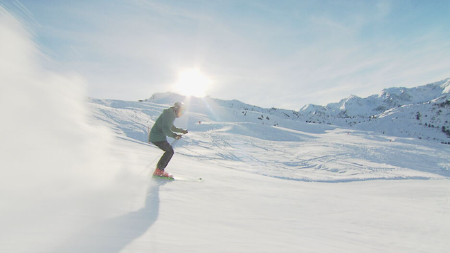 Raich fährt allein die Piste hinunter, die Sonne strahlt hell, die Berge sind weiß, Schnee wirbelt auf