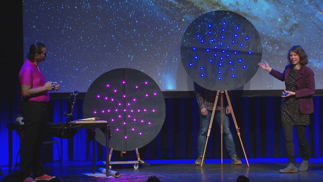 Bühne: links: Martin Puntigam in neonpinkem engen T-Shirt, neben ihm zwei schwarze Gebilde aus Pappe mit leuchtenden Punkten, Grützbauch deutet darauf und erklärt