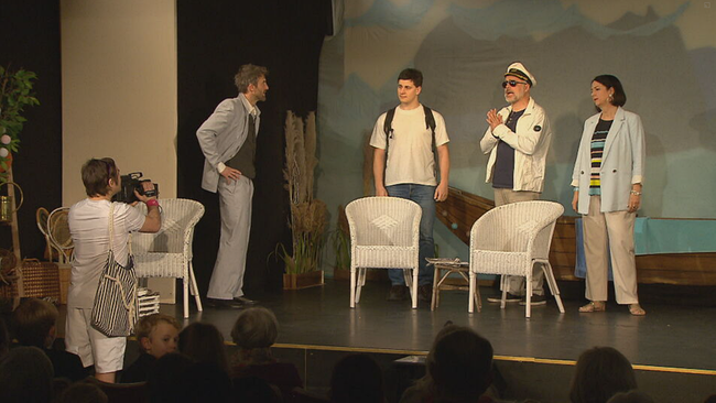 Bühnenbild: Strandsituation, zwei weiße Stühle, ein Kamermann mit Strandtasche filmt, eine Frau und drei Männer auf der Bühne