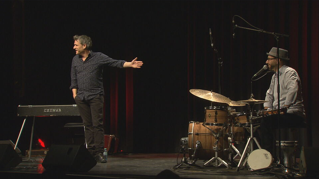 Spörk auf der Bühne, deutet mit einer Hand auf den Kollegen Alberto Lovison am Schlagzeug, Blick zum Publikum