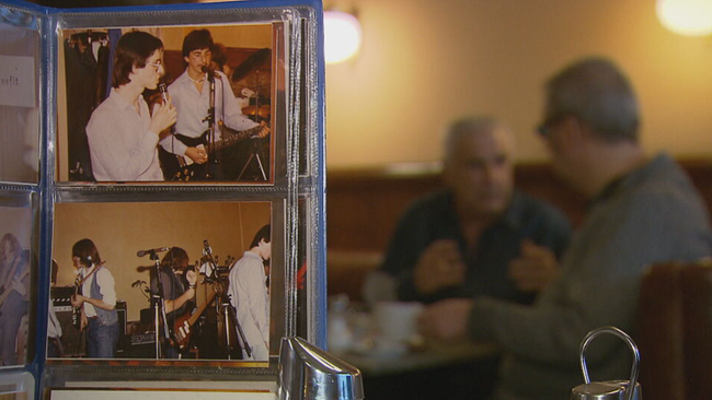 Im Vordergrund: Ein Bilderalbum mit Fotos eines Konzerts aus den 80ern, im Hintergrund Krankl und Schreiber am Tisch