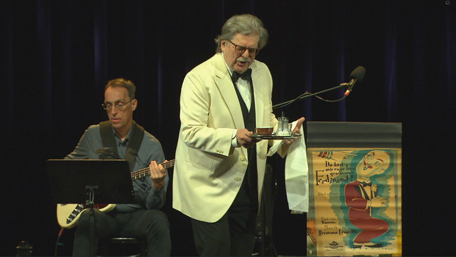 © ORF2/Seitenblicke Erwin Steinhauer im Anzug mit Weste und Fliege und wießem Sakko, auf dem Arm eine Stoffserviette und in der Hand ein Tablett mit einem Espresso und Zucker, singend. Im Hintergrund ein Leopoldi Plakat und der Gitarrist