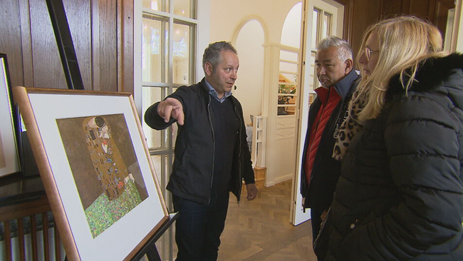 Alakus deutet mit dem Finger erklärend auf ein Replikat von Klimts "Der Kuss", vor ihm zwei BesucherInnen, die zuhören