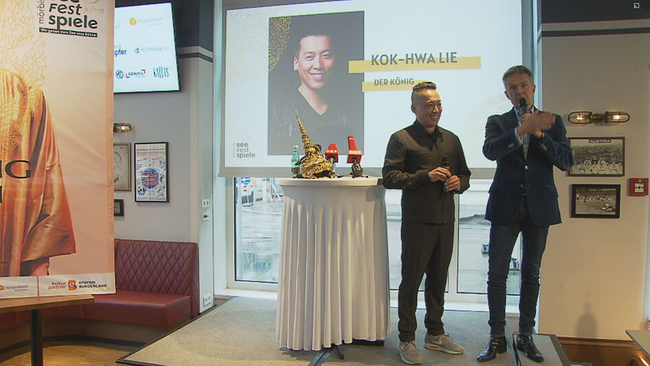 Pressekonferenz: Kok-Hwa Lie und Haider auf einer kleinen Bühne, auf dem Tisch die Krone, im Hintergrund eine Slideshow mit Foto von Lie