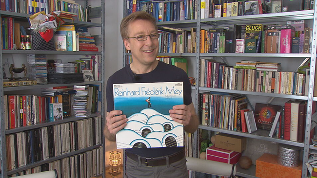 Müller steht in der Ecke, links und rechts zwei groé Regale mit Schallplatten, Büchern und Zeug, er steht mit einer blauen "Reinhard Frédérik Mey" Platte davor und strahlt