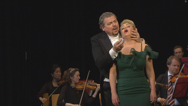 Michael Schade im Anzug hält singend Daniela Fally in grünem Kleid im Arm, die zu sterben scheint. Orchester im Hintergrund