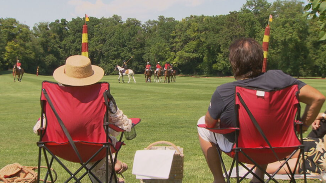 CamperInnen-Pärchen in roten Aufklappstühlen, zwischen Ihnen Picknickkorb, in der Entfernung Pferde beim Polospiel