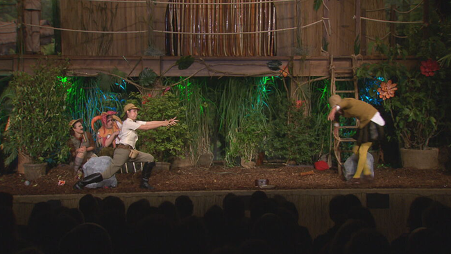 Bühnenbild Dschungel: Vier Personen sind zu sehen, eine Frau im Soldatenkostüm tut so als würde sie schießen