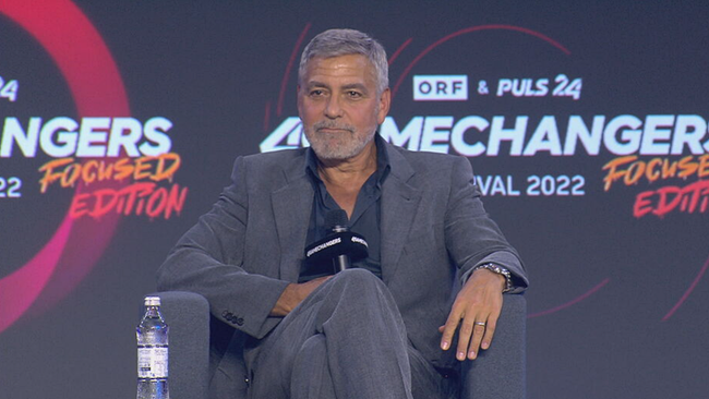 Clooney sitzt in dunkelgraum Anzug gemütlich auf der Bühne