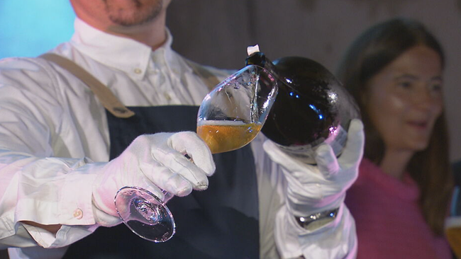 Kellner in weißem Hemd, mit weißen Handschuhen und Schürze schenkt aus der Flasche das Bier in ein Glas ein