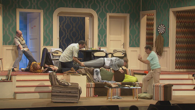 Fünf Männer raufen auf der Bühne im 60er Set eines Wohnzimmers