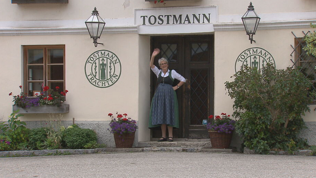 Tostmann steht winkend vor der Tür eines renovierten Bauernhauses, über der Tür steht "Tostmann", links und rechts der Aufdruck "Tpstmann Trachten"
