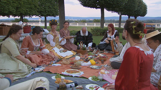 mindestens 8 Personen sitzen in barocker Kostümierung auf Picknickdecken mit Wein und Obst in der Mitte