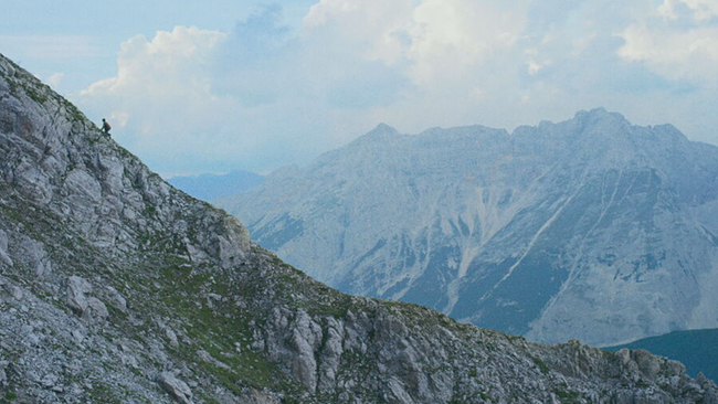 Elias klettert einen massiven Berg hinauf, im Hintergrund ein See und weitere Gebirge