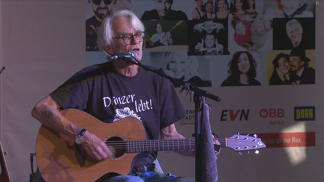 Ulli Bäer sitzt mit Gitarre auf der Bühne. Er trägt ein "Danter lebt" T-Shirt 