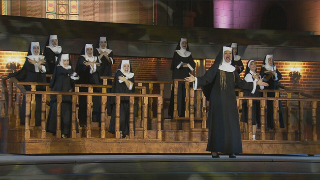 Musicaldarsteller in Nonnen Kostümen stehen auf der Bühne die Chorreihen einer Kirche andeutet 