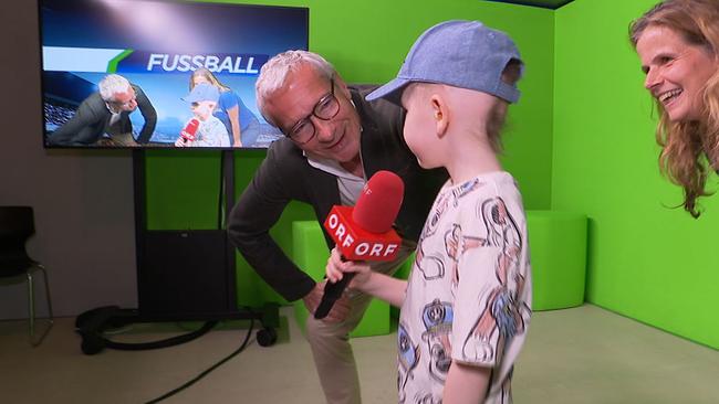 Ein Kind mit schütterem Haar unter einer Kappe hält ein ORF-Mikrofon in der Hand.