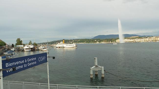 Zu sehen sind eine große Wasserfläche, ein Stadt im Hintergrund und ein Schild, auf dem auf Französisch und Englisch "Willkommen in Genf!" steht.