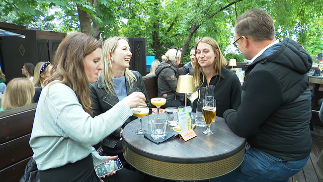 Vier Menschen stehen im Freien rund um einen Stehtisch, auf dem sich Cocktails und andere Getränke befinden.