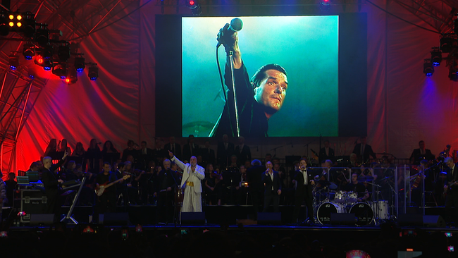 Auf einer Bühne ist eine Leinwand zu sehen, von der aus der verstorbene Falco zum Publikum von 2024 singt.