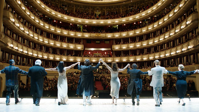 Eine Gruppe von Darstellern steht mit dem Rücken zur Kamera auf einer Opernbühne und verneigt sich in Richtung eines vollen Zuschauerraums.