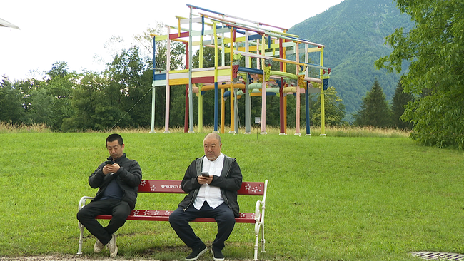 Zwei Männer sitzen auf einer Parkbank vor einem überdimensionalem Kunstwerk im Hintergrund.