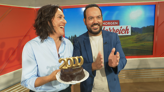 Eine Frau und ein Mann halten lächeld eine Geburtstagstorte mit der Zahl "2000" darauf.