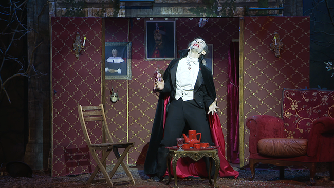 Ein mit rotem Umhang, bleicher Maske und Vampirzähnen kostümierter Darsteller spielt auf einer Theaterbühne.  