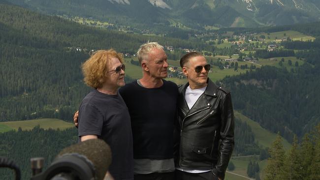 Drei mittelalte Herren stehen auf einer Wiese, hinter ihnen ist eine grüne Berglandschaft zu sehen.