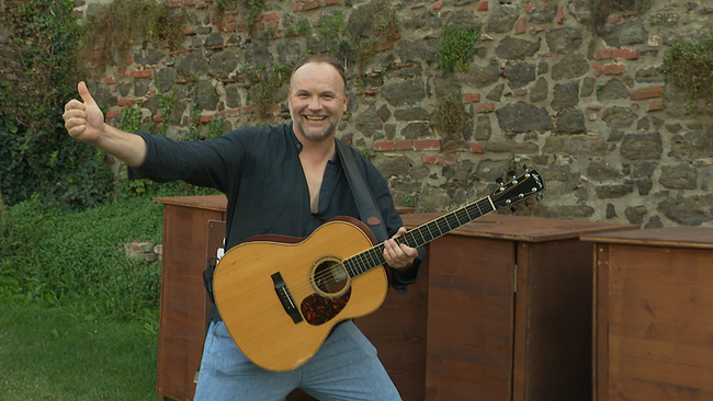 Ein Mann mit Halbglatze und Gitarre lächelt in die Kamera.