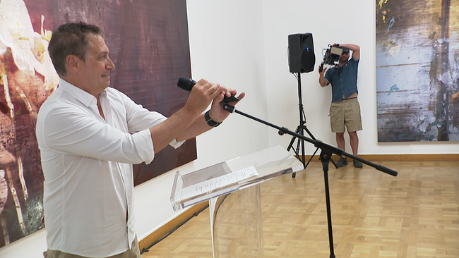 © ORF / Seitenblicke Ein Mann, der in einem Museum vor großformatigen Bildern steht, richtet sich sein Mikrofon ein.