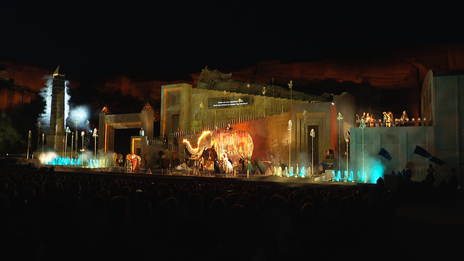 Auf einer Bühne, hinter der ein gigantischer Steinbruch zu sehen ist, tummeln sich Tänzer mit Fackeln neben einem hölzernen Elefanten. 