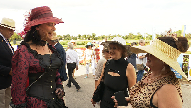 Zu sehen sind einige Damen, die am Rande eine Pferderennbahn stehen und alle sehr opulente Hüte tragen.