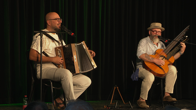 Zwei Musiker  - der eine mit Zieharmonika und der andere mit Gitarrre - sitzen auf einer Bühne und spielen. 