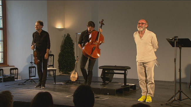 Schauspieler Karl Markovics steht zusammen mit Matthias Bartolomey (Violoncello) und Klemens Bittmann (Violine und Mandola) und begrüßt das Publikum 