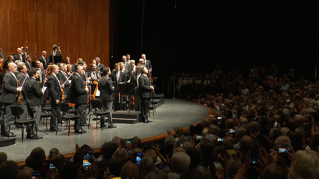 Ein Dirigent steht in einer Konzerthalle vor seinem Orchester.
