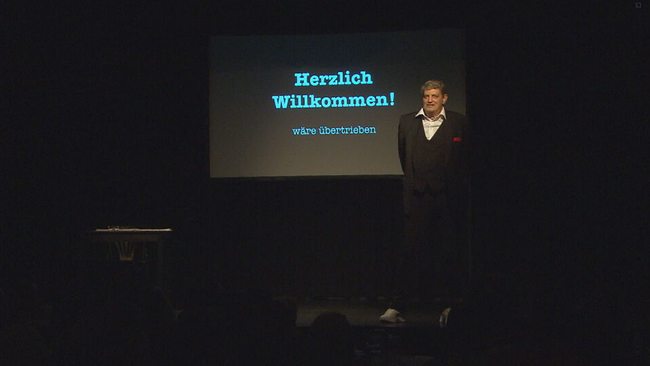 Dieter Chemelar steht auf der Bühne der Kulisse in Wien. Im Hintergrund ist eine Leinwand mit der Aufschrift: Herlich Willkommen (Wäre übertrieben) 