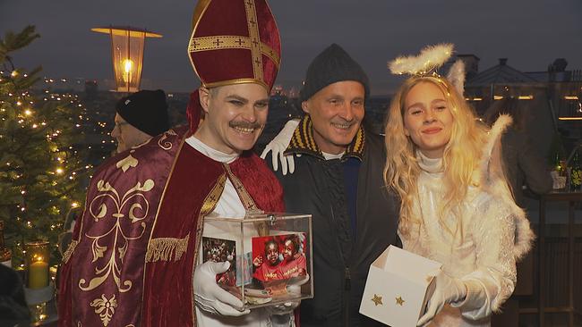 Ein als Nikolaus verkleideter Mann mit einer Spendenbox, ein Mann mit einer Wintermütze und eine als Christkind verkleidete Frau lächeln in die Kamera.