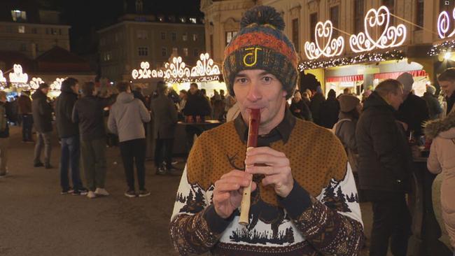 Ein Mann in einer Winterhaube steht auf einem Weihnachtsmarkt und spielt Blockflöte.