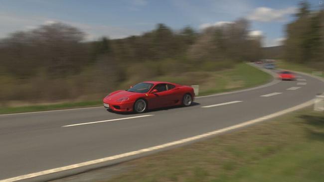 Roter Ferrari fährt auf der Straße, dahinter unscharf andere Sportwagen 