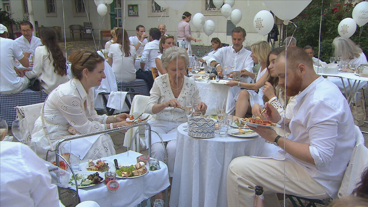 Menschen ganz in weiß gekleidet picknicken an einem Tisch. Rund um sie schweben weiße Luftballons 