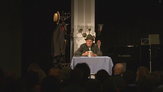 Robert Meyer sitzt im Trachtenoutfit auf der Bühne vor einem Tisch mit blau/weiß karriertem Tischtuch. In der Hand hat er einen Maßkrug