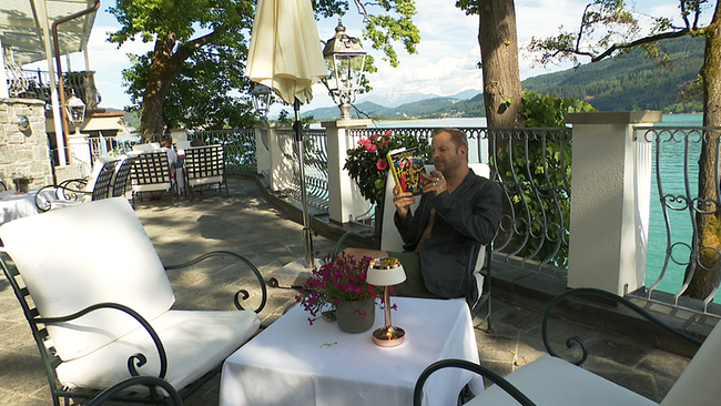 © ORF / Seitenblicke Philipp Hochmair sitzt an einem Tisch am See und liest sein Buch