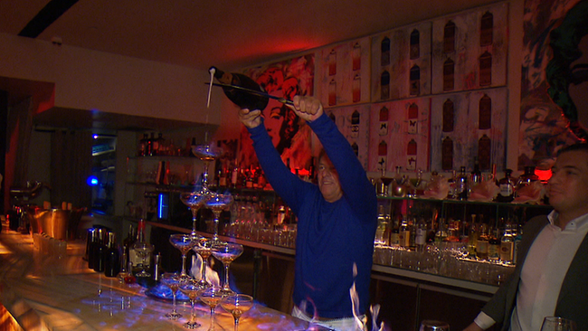 © ORF / Seitenblicke Ein Mann köpft hinter einer Bar eine Prosecco Flasche und befüllt damit in flammen stehende Prosecco Gläser.